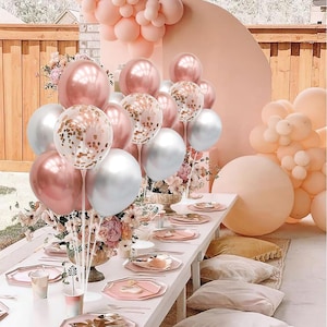 May include: A festive balloon centerpiece featuring a mix of metallic rose gold and silver balloons, including clear balloons filled with rose gold confetti. The arrangement is displayed on a long table set with plates and floral decorations.