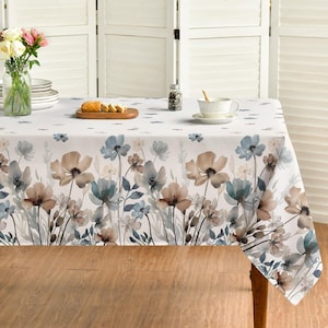 May include: A rectangular tablecloth featuring a white base with a watercolor-style floral print in shades of blue, beige, and brown. The pattern displays various wildflowers and stems extending upward from the bottom edge across the fabric surface.