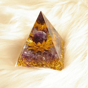 Peut inclure: Une pièce d'art en résine transparente en forme de pyramide avec une pierre violette et des accents floraux dorés. La pyramide est remplie de paillettes violettes et dorées, créant un objet unique et décoratif.