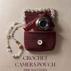 May include: A dark red crochet camera pouch with a cream-colored scalloped edge and a single white button closure. A red digital camera is tucked inside the pouch. Beside it lies a beaded lanyard featuring clear beads, small white starfish, and shells.