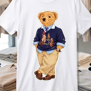 May include: A white short-sleeved t-shirt featuring a graphic print of a teddy bear wearing a navy blue sweater with a polo player design, a light blue collared shirt, a tie, tan trousers, and brown loafers, standing with hands in pockets.