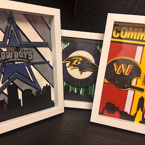 May include: Three white-framed shadow box art pieces featuring NFL team themes. The left frame displays a blue star and the text Cowboys. The center frame shows a purple raven logo. The right frame features two helmets and the text Commanders.