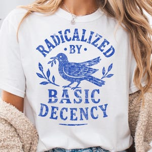 以下が含まれることがあります： 白のTシャツに青いグラフィック。グラフィックは、オリーブの枝を運ぶ鳩と、周りに「RADICALIZED BY BASIC DECENCY」という言葉がかすれたフォントで描かれています。このシャツはカジュアルウェアです。