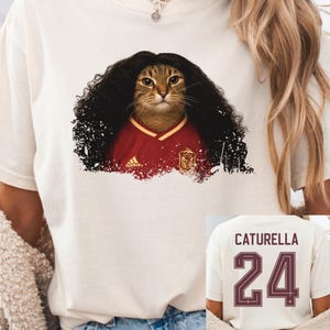Op de afbeelding: Een off-white t-shirt met een afbeelding van een bruine cyperse kat met lang donker krullend haar in een rood voetbalshirt. De achterkant van het shirt toont de tekst CATURELLA boven het nummer 24 in een donkerrood sportief lettertype.