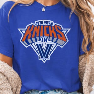 Op de afbeelding: Een koningsblauw T-shirt met korte mouwen en een verweerde grafische print op de borst. Het ontwerp toont de tekst NEW YORK KNICKS IN V in oranje, wit en lichtblauw, gerangschikt in een driehoekige vorm met een grote V aan de onderkant.