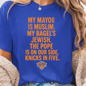 Op de afbeelding: Een koningsblauw t-shirt met korte mouwen en oranje verweerde tekst: MY MAYOR IS MUSLIM. MY BAGEL'S JEWISH. THE POPE IS ON OUR SIDE, KNICKS IN FIVE. Onder de tekst staat het officiële logo van het basketbalteam de New York Knicks.
