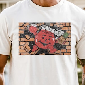 Pode incluir: Uma t-shirt de manga curta cor de creme com uma impressão gráfica de uma personagem em forma de jarro vermelho a atravessar uma parede de tijolos. A personagem segura um ananás e um balão de fala com o texto DAT BIH GAH.