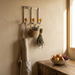 Peut inclure: Un porte-manteau mural en métal avec trois crochets en verre ambré. Un tablier beige, un petit panier en osier et des herbes séchées sont suspendus aux crochets. Un bol de fruits est posé sur une surface en bois.