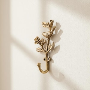 May include: A brass-toned metal wall hook featuring a sculpted design of a small bird perched on a branch with leaves and flowers. The hook has a curved bottom and is attached to the wall with two visible screws at the center of the structure.