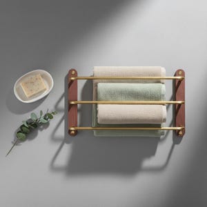 May include: A wooden towel rack with three brass bars holds three folded towels in beige and green. A white soap dish with a bar of soap and a sprig of greenery are to the left.