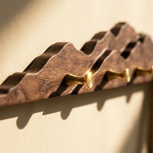 May include: A wooden wall-mounted coat rack with a mountain-like design. The rack is made of dark brown wood and features several brass-colored knobs for hanging items. The design casts interesting shadows on the wall.