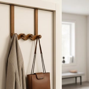 Könnte beinhalten: Eine hölzerne Türgarderobe mit wellenförmigem Design und vier abgerundeten Haken. Die Garderobe ist oben an der Tür eingehängt und hält links einen beigen Mantel sowie rechts eine braune Ledertasche mit Kettenriemen an den Haken.
