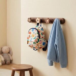 Può includere: Un attaccapanni a muro in legno con pioli a forma di fiore. Uno zaino colorato con un motivo di animali dei cartoni animati è appeso a un piolo e un maglione azzurro è appeso a un altro. Sotto c'è uno sgabello di legno.
