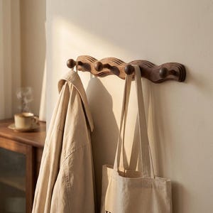 May include: A wooden coat rack with five pegs, mounted on a wall. The rack is made of dark brown wood and has a wavy design. A beige jacket and a canvas tote bag hang from the pegs. The wall is painted a light beige colour.