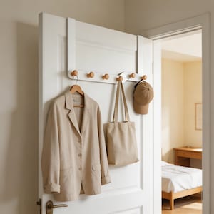 May include: A white over-the-door organiser with six wooden knobs. A beige blazer hangs on a wooden hanger, a beige tote bag, and a brown baseball cap are also displayed. The door is white, and the room beyond is softly lit.