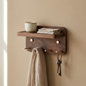 May include: A dark wood wall-mounted shelf with a small shelf, brass hooks, and brass accents. A beige scarf hangs from a hook, and keys hang from a leather loop. A ceramic cup and folded cloth sit on the shelf.