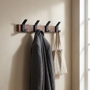 May include: A wooden wall-mounted coat rack with four black metal hooks. A gray coat and a beige tote bag hang from the hooks. The rack is mounted on a light-colored wall near a window.