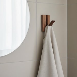 Op de afbeelding: Een houten wandhaak met een handdoek eraan. De haak bestaat uit twee verticale stukken hout met een horizontaal stuk dat uitsteekt. Een lichtgekleurde handdoek is over de haak gedrapeerd. Een ronde spiegel is zichtbaar op de achtergrond.