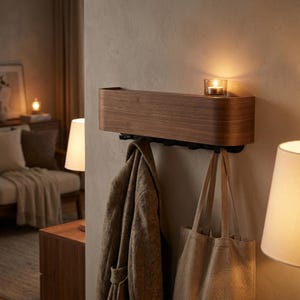 Puede incluir: Estante de pared de madera con borde frontal curvado y ganchos debajo. Una vela encendida está sobre el estante. Un abrigo y un bolso de mano cuelgan de los ganchos. El estante es de madera oscura y está montado en una pared de color neutro.