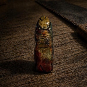 May include: A hand-carved wooden sculpture with a textured surface, featuring a blend of colors including red, green, and gold. The sculpture is set against a wooden background, with a saw in the background.