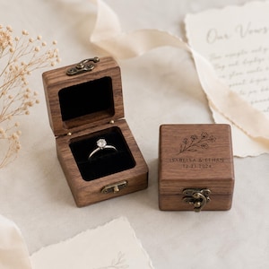 May include: Two wooden ring boxes, one open with a diamond ring, the other closed. The closed box has floral engraving and the names "ISABELLA & ETHAN" with the date "12.21.2024". Both boxes have brass-colored clasps.