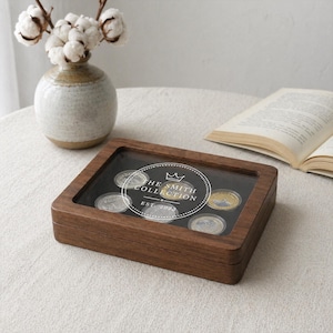 May include: A wooden coin display case with a clear top, showcasing several coins. The case has a dark wood finish and features the text "THE SMITH COLLECTION EST. 2023" on the inside. A vase with cotton stems and a book are in the background.