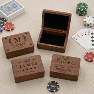 Op de afbeelding: Vier houten dozen met gepersonaliseerde gravures, inclusief namen en data, worden getoond met speelkaarten en pokerchips. Eén doos is open en onthult een zwart interieur. De dozen hebben speelkaartsymbolen en kroonontwerpen.