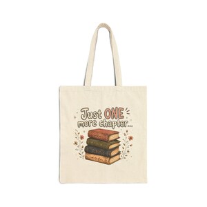 Op de afbeelding: Een beige canvas draagtas met een grafische print van een stapel van vier boeken met bloemaccenten aan de zijkanten. Boven de boeken staat de tekst Just ONE more chapter in een gestileerd lettertype. De tas heeft twee lange schouderbanden.