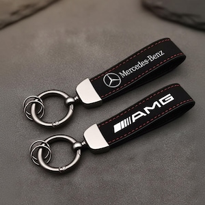 Peut inclure: Deux porte-clés en cuir noir avec des anneaux argentés. L'un porte le logo et le texte Mercedes-Benz en blanc, l'autre le logo et le texte AMG en blanc. Les deux ont des coutures rouges.