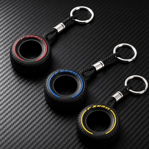 Pode incluir: Três porta-chaves com pneus de borracha preta em miniatura com padrão de rasto. Cada pneu tem um anel colorido e o texto P ZERO impresso na parede lateral. As cores são vermelho, azul e amarelo. Cada pneu liga-se a um anel de metal.