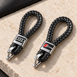 Peut inclure: Deux porte-clés en cuir tressé noir avec des éléments métalliques argentés. L'un des porte-clés présente le logo Audi à quatre anneaux en blanc sur fond noir, tandis que l'autre affiche le logo S line rouge et blanc sur fond noir.