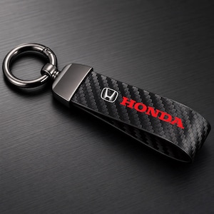 Peut inclure: Porte-clés en fibre de carbone noire avec un anneau et un fermoir argentés. Le porte-clés présente le logo Honda en blanc et le mot "HONDA" en rouge. Le fond est une surface sombre et texturée.