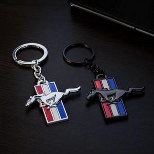 Peut inclure: Deux porte-clés en métal représentant un emblème de cheval au galop sur une barre rectangulaire verticale avec des bandes rouges, blanches et bleues. L'un a une finition argentée polie avec un anneau argenté, l'autre une finition noire mate.