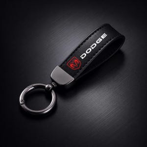 May include: Black leather keychain with the word "DODGE" in white and a red ram logo. The keychain has a silver-colored metal clasp and loop for attaching keys. The background is a dark, textured surface.