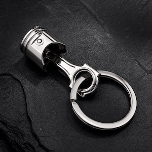 May include: A silver-coloured metal keyring featuring a miniature engine piston and connecting rod assembly. The cylindrical piston head is attached to a sturdy metal ring, creating a functional accessory with a polished, reflective finish.