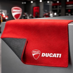 Può includere: Un tappeto Ducati rosso e grigio con il logo Ducati in bianco. Il tappeto è rettangolare e ha una consistenza morbida simile al feltro. Il logo Ducati è uno scudo bianco con la scritta "Ducati" in bianco.