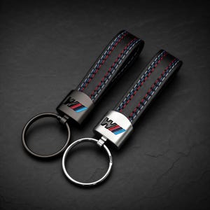 Op de afbeelding: Twee sleutelhangers met zwarte leren bandjes en blauwe, witte en rode stiksels. Elk bandje is bevestigd aan een metalen connector met de letter W en blauwe en rode strepen. Eén sleutelhanger heeft een matzwarte ring, de andere een zilveren ring.