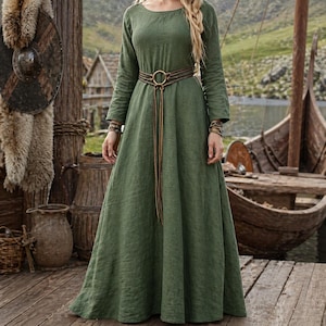 May include: An olive green long-sleeve dress with a round neckline and a flowing skirt. The dress is cinched at the waist with a brown leather belt and a long, matching tie. The dress is part of a historical or medieval themed collection.