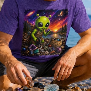 May include: A heather purple t-shirt featuring a graphic print of a green alien character collecting colorful rocks on a beach. The alien wears a vest and holds a small stone, with a bag labeled ROCKS OUT OF THIS WORLD nearby under a starry sky.