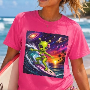 May include: A bright pink short-sleeved t-shirt featuring a square graphic print of a green alien surfing on a wave. The illustration includes a sunset, palm trees, planets, and a galaxy in the background. The alien wears a bikini and a flower.
