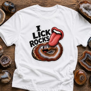 以下が含まれることがあります： 白いTシャツに、茶色と白のジオード（晶洞石）のキャラクターが大きな赤い舌で舐められているグラフィックが描かれています。ジオードの上には「I LICK ROCKS」という太い黒文字がプリントされています。シャツの周りには、磨かれた様々なメノウ石と黒い虫眼鏡が配置されています。