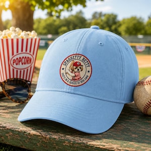 May include: A light blue baseball cap featuring a circular graphic on the front. The graphic depicts a dog wearing a red baseball cap and holding a bat, surrounded by the text "SPAGHETTY BETTY" and "SHE'S IN A LEAGUE OF HER OWN".