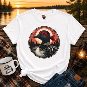 May include: A white t-shirt featuring a circular graphic of a loon wearing a red bandana and a silver necklace with a Minnesota-shaped pendant. The background of the graphic shows a sunset over a lake with silhouetted pine trees and birds.