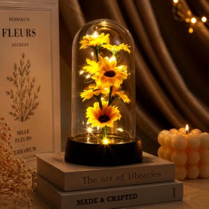 May include: A glass cloche displaying yellow sunflowers illuminated by fairy lights. The cloche is set on a black base, placed on two books with the titles "The art of libraries" and "MADE & CRAFTED". A lit candle is in the background.