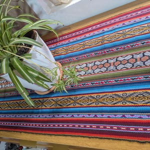 May include: A rectangular table runner featuring a vibrant woven pattern with horizontal stripes in shades of blue, red, orange, green, and purple. The fabric displays intricate geometric diamond and honeycomb motifs throughout the colorful textile design.