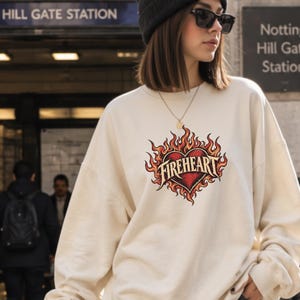 May include: A sand-coloured crewneck sweatshirt featuring a graphic print of a red heart surrounded by orange and yellow flames with the text "FIREHEART" written across the centre in a bold, stylised font. The garment has long sleeves and ribbed cuffs.