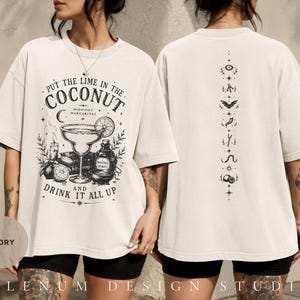Op de afbeelding: Een ivoorkleurig t-shirt met een zwarte grafische print. De voorkant toont een margaritaglas, limoenschijfjes en de tekst "Put the lime in the coconut and drink it all up". De achterkant bevat een verticale kolom met kleine mystieke symbolen.