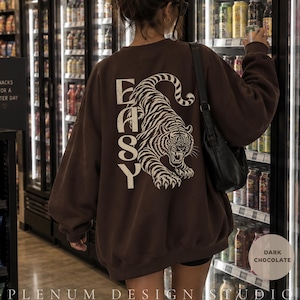 Op de afbeelding: Een donkerbruin sweatshirt met ronde hals en een grote crèmekleurige afbeelding op de achterkant. Het ontwerp bevat een gedetailleerde illustratie van een tijger in een springende houding naast de verticale tekst "EASY" in een verweerd bloklettertype.