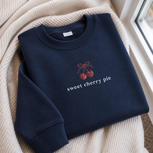Puede incluir: Una sudadera azul marino de cuello redondo con un bordado de dos cerezas rojas atadas con un lazo y el texto blanco "sweet cherry pie" centrado en el pecho. La prenda tiene mangas largas con puños acanalados y un cuello acanalado.