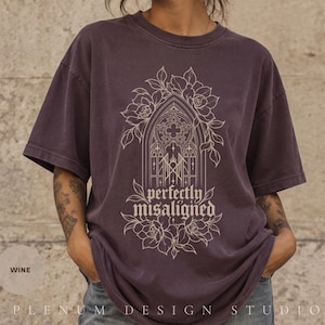Pode incluir: Uma t-shirt de manga curta cor de vinho com um gráfico de linhas bege de uma janela gótica arqueada rodeada por elementos florais. O texto "perfectly misaligned" está impresso numa fonte gótica decorativa dentro do design.